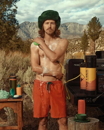 Man showers outdoors near a mountain range, wearing orange swim trunks and a green hat. Camping equipment is near him. 